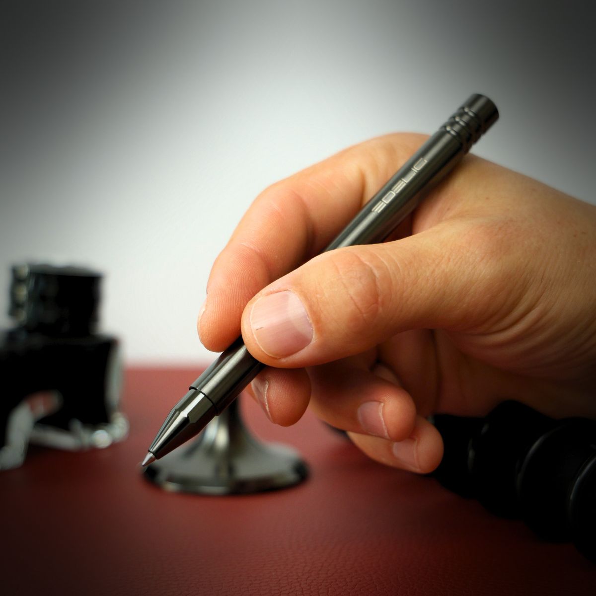 Eine männliche Hand hält einen eleganten Tintenroller (Schaft) aus Edelstahl in DCL-Anthrazit. Im Hintergrund, unscharf, sind der Standfuß des Tintenrollers sowie ein Tintenfass mit schwarzer Tinte zu erkennen. Dieses Schreibgerät verkörpert ein spürbares Gewicht, eine metallische Haptik und ein elegantes Schreibgefühl.