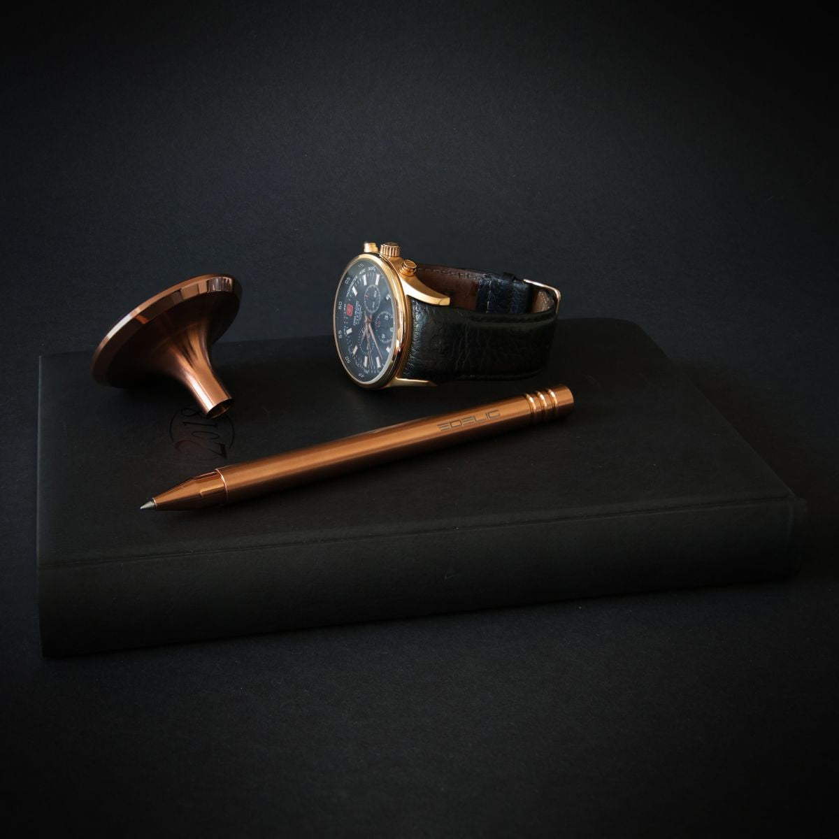 Eleganter Tintenroller V4INK in Kupferoptik präsentiert sich stilvoll als Accessoire neben einer hellkupferfarbenen Schweizer Uhr auf einem schwarzen Notizbuch. Diese Komposition verkörpert die Eleganz in Formgebung und der Detailverarbeitung des Schreibgeräts.