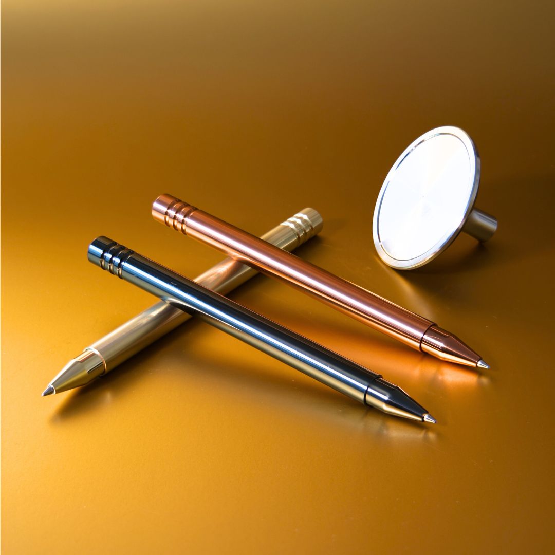 Auf einem goldfarbenen Hintergrund liegen drei luxuriöse Tintenroller in edlen Metall- Finishes: DLC- Anthrazit, PVD- Kupfer und PVD- Kupfer. Die Schreibgeräte zeichnen sich durch ihre elegante PVD- Beschichtung aus, die ihnen eine besonders widerstandsfähige und exklusive Oberfläche verleiht. Im Hintergrund ist ein glänzendes Standfuß aus purem Edelstahl als Basis zu sehen.