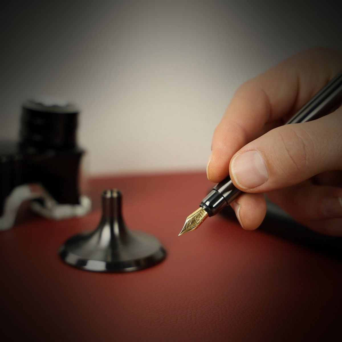 Eine männliche Hand hält einen eleganten Füllfederhalter (Schaft) aus Edelstahl in DCL-Anthrazit. Im Hintergrund, unscharf, sind der Standfuß des Füllfederhalters sowie ein Tintenfass mit schwarzer Tinte zu erkennen. Dieses Schreibgerät verkörpert ein spürbares Gewicht, eine metallische Haptik und ein elegantes Schreibgefühl.