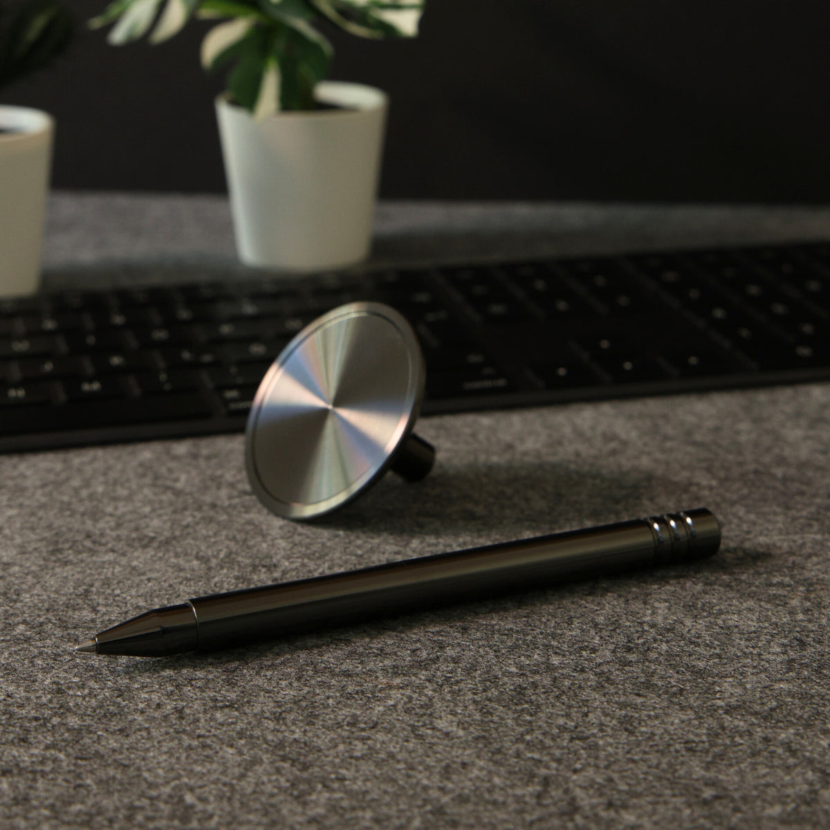 Edles Büro-Setup mit Tintenroller in DLC-_Anthrazit. Stiftschaft und Standfuß getrennt liegend. Im Hintergrund eine schwarze Tastatur auf Filzunterlage. Zwei Zimmerpflanzen in weißen Töpfen sorgen für eine stilvolle, minimalistische Atmosphäre.