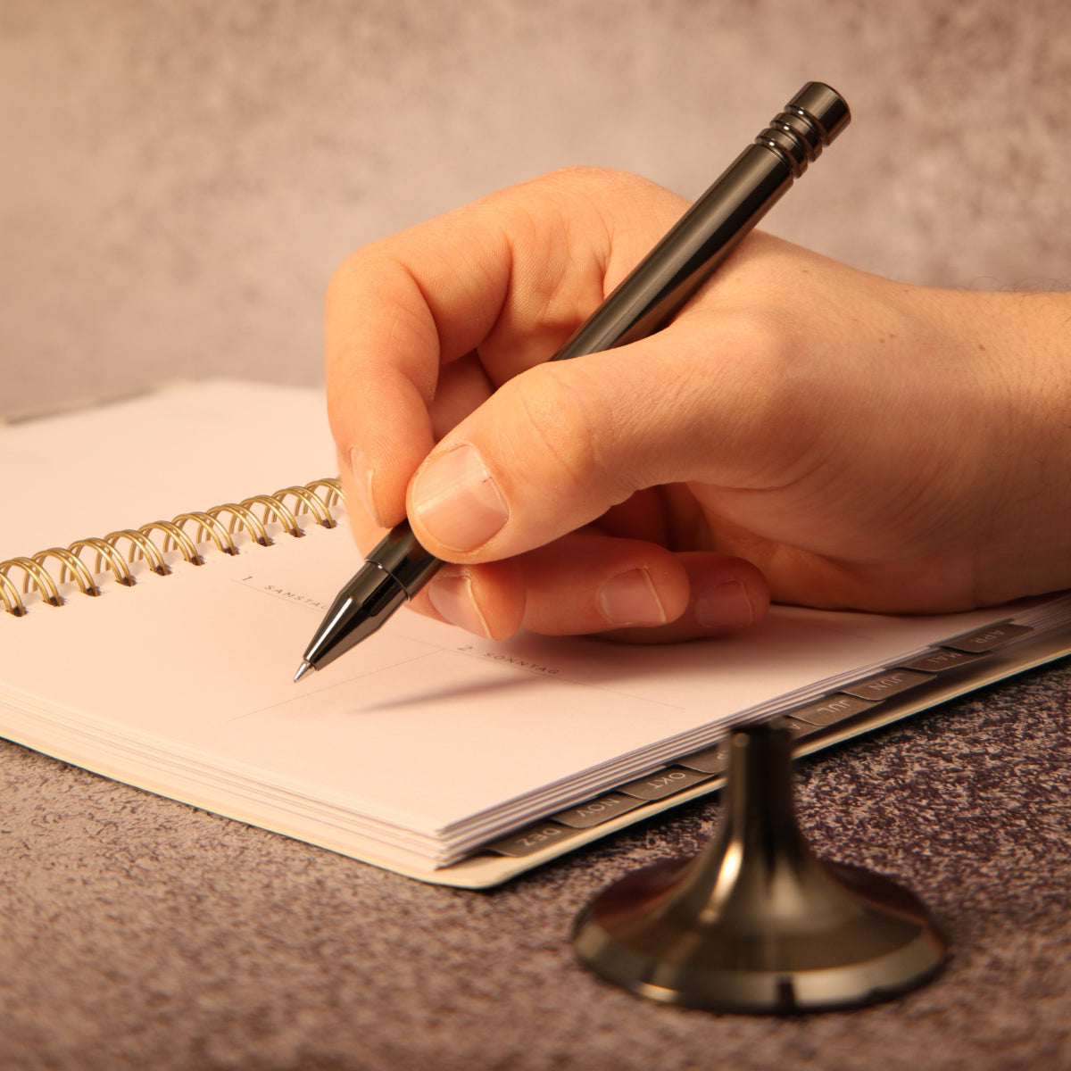 Eleganter, zweigeteilter Tintenroller aus DLC- Anthrazit, bestehend aus einem Schaft und einem Standfuß. Das Schreibgerät liegt harmonisch in der Hand, die auf einem Notizbuch ruht. Im Vordergrund des Bildes ist der Standfuß sichtbar.