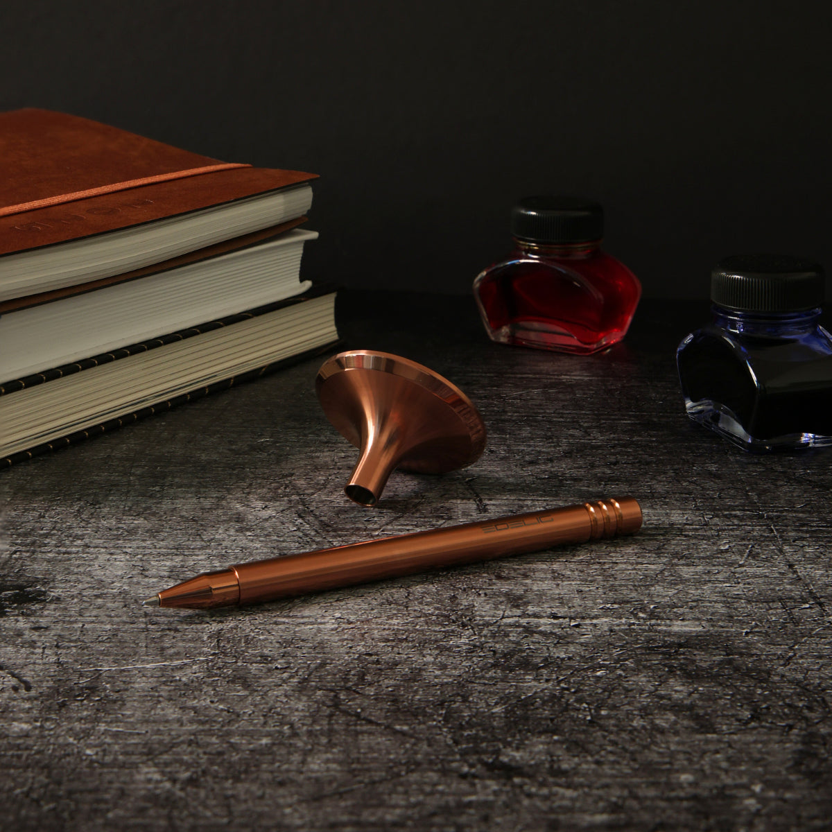 Ein elegantes Setup mit einem Tintenroller in PVD-Kupfer in einer abgedunkelten Umgebung. Im Hintergrund sorgen drei Bücher und zwei Fläschchen mit blauer und roter Tinte für eine stilvolle, romantische Atmosphäre.