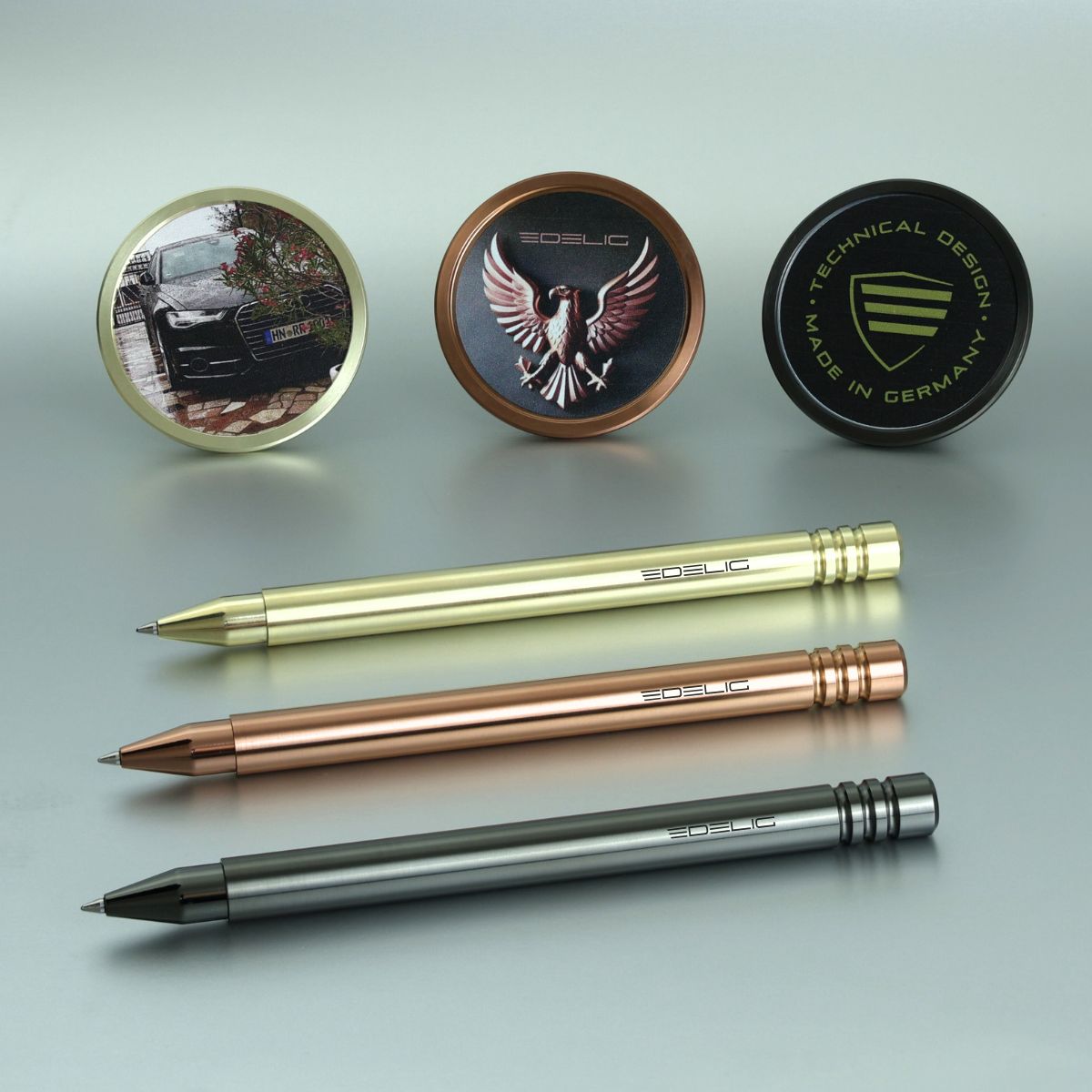 Personalisiertes Schreibgerät mit einem individuellen UV- Direktdruck. Auf dem Bild sind die V4INK- Modelle in drei Farben und Grafiken dargestellt, wobei Standfuß und Schaft nebeneinander liegen. Das V4INK in PVD- Hellgold zeigt die Grafik des Audi A6, während das V4INK in PVD- Kupfer mit der Grafik eines Adlers sowie dem EDELIG-Logo versehen ist. Das V4INK in DLC- Anthrazit präsentiert das Wappen von EDELIG mit dem runden Schriftzug „technical design - made in Germany“.