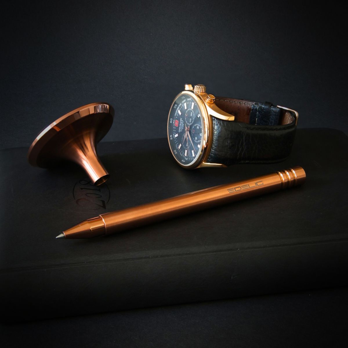 Eleganter Tintenroller V4INK in Kupferoptik präsentiert sich stilvoll als Accessoire neben einer hellkupferfarbenen Schweizer Uhr auf einem schwarzen Notizbuch. Diese Komposition verkörpert die Eleganz in Formgebung und der Detailverarbeitung des Schreibgeräts.